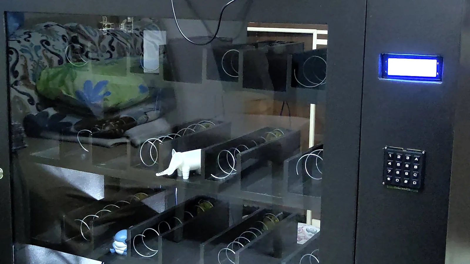 RoboMed eczane otomatının ön yüzü: camlı kapak arkasında 16 spiral hazne, yanan LCD ekran ve tuş takımı